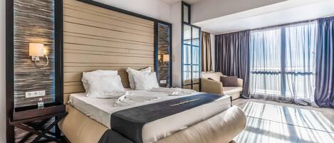A modern hotel room with a large bed, a sofa, a desk with a lamp, and a large window with curtains.
