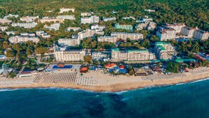 An aerial view of a coastal resort with multiple hotel buildings, a beach with sun loungers, and a clear blue sea. - Prestige Deluxe Hotel & Aquapark Club - All Inclusive (Golden Sands)