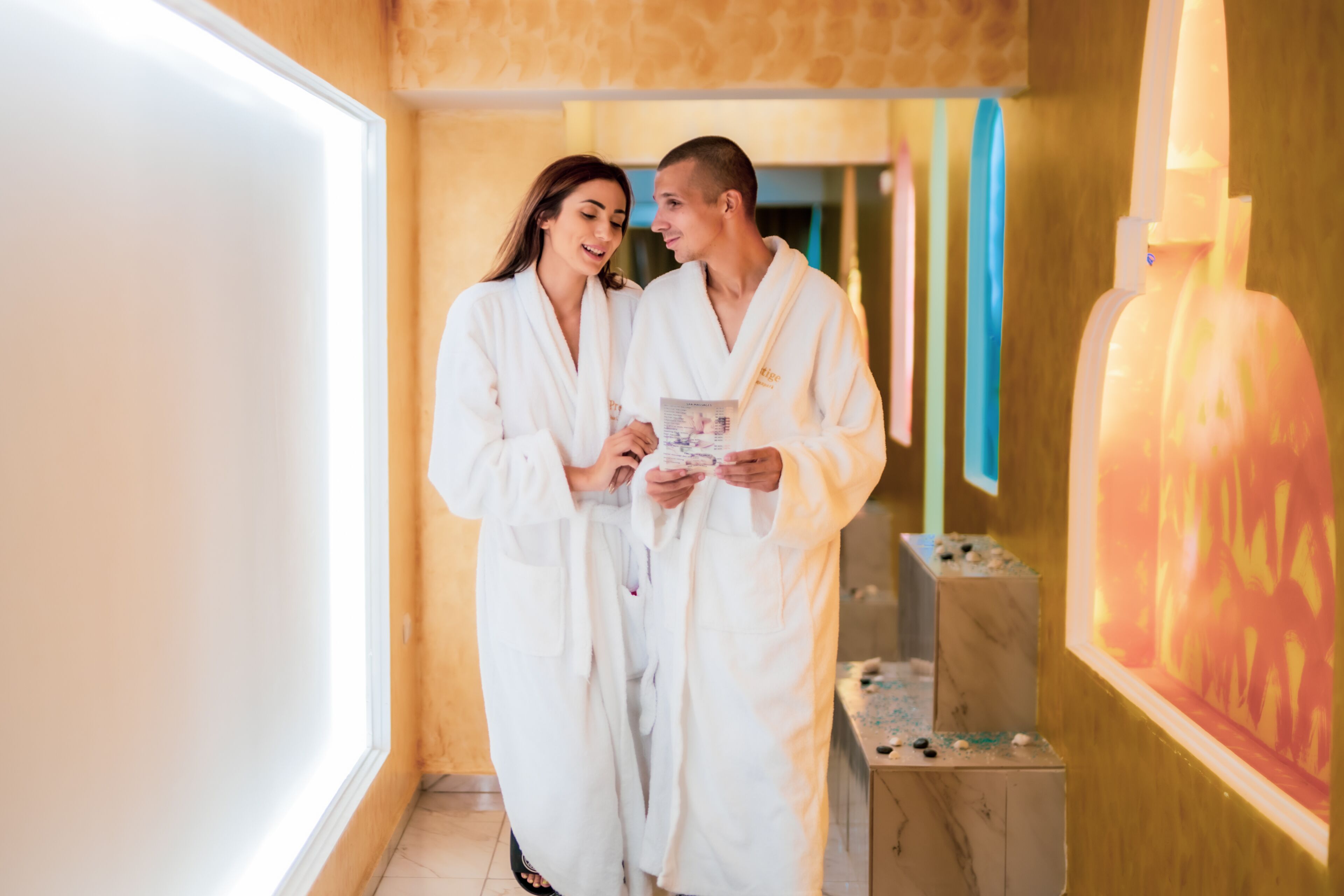 Una pareja con batas blancas en un ambiente de spa, con una mesa de masajes y una silla de masajes a la vista.