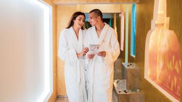 Una pareja con batas blancas en un ambiente de spa, con una mesa de masajes y una silla de masajes a la vista.