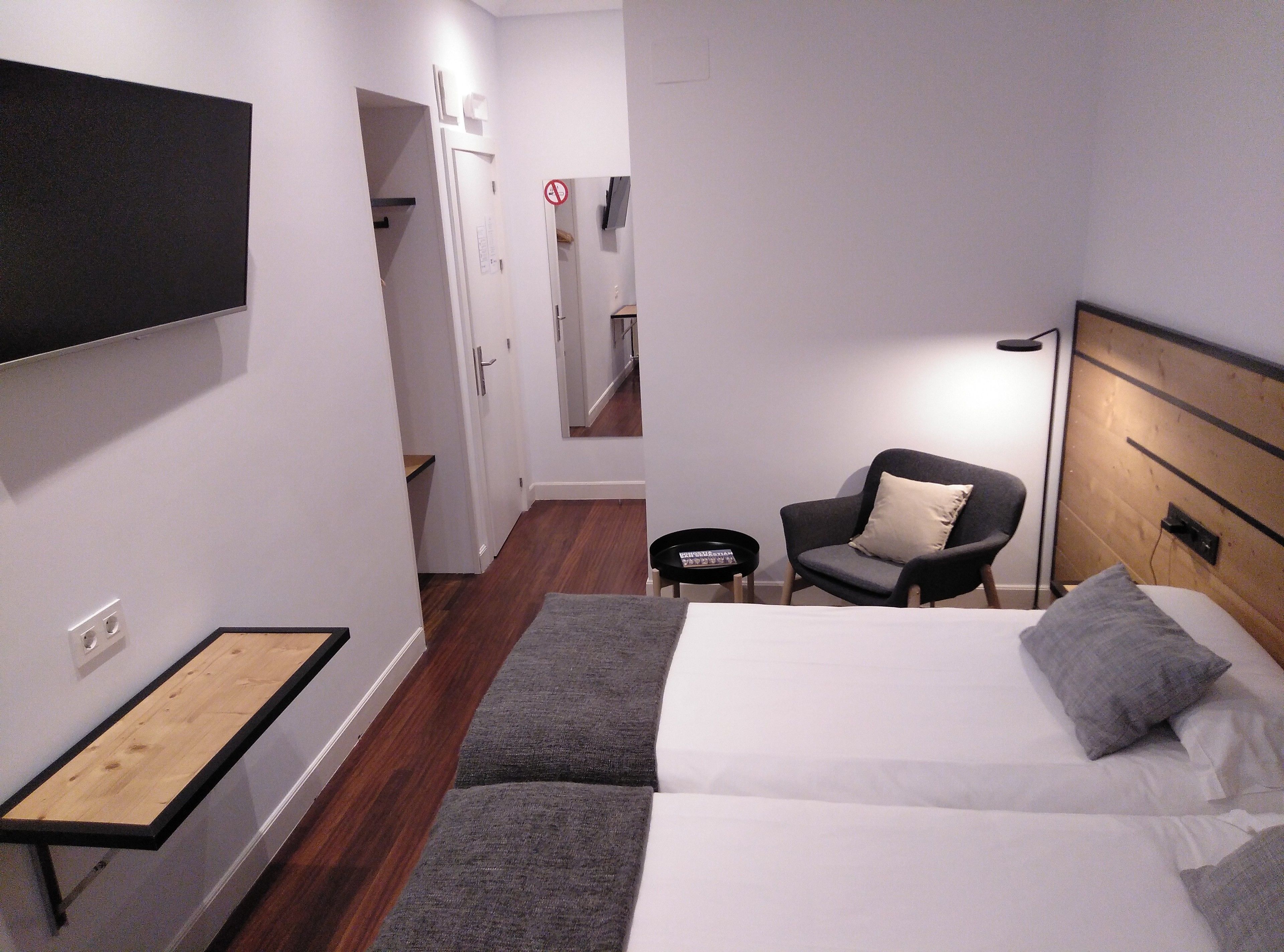 A hotel room with a bed, a chair, a small table, a TV mounted on the wall, and a wooden floor.