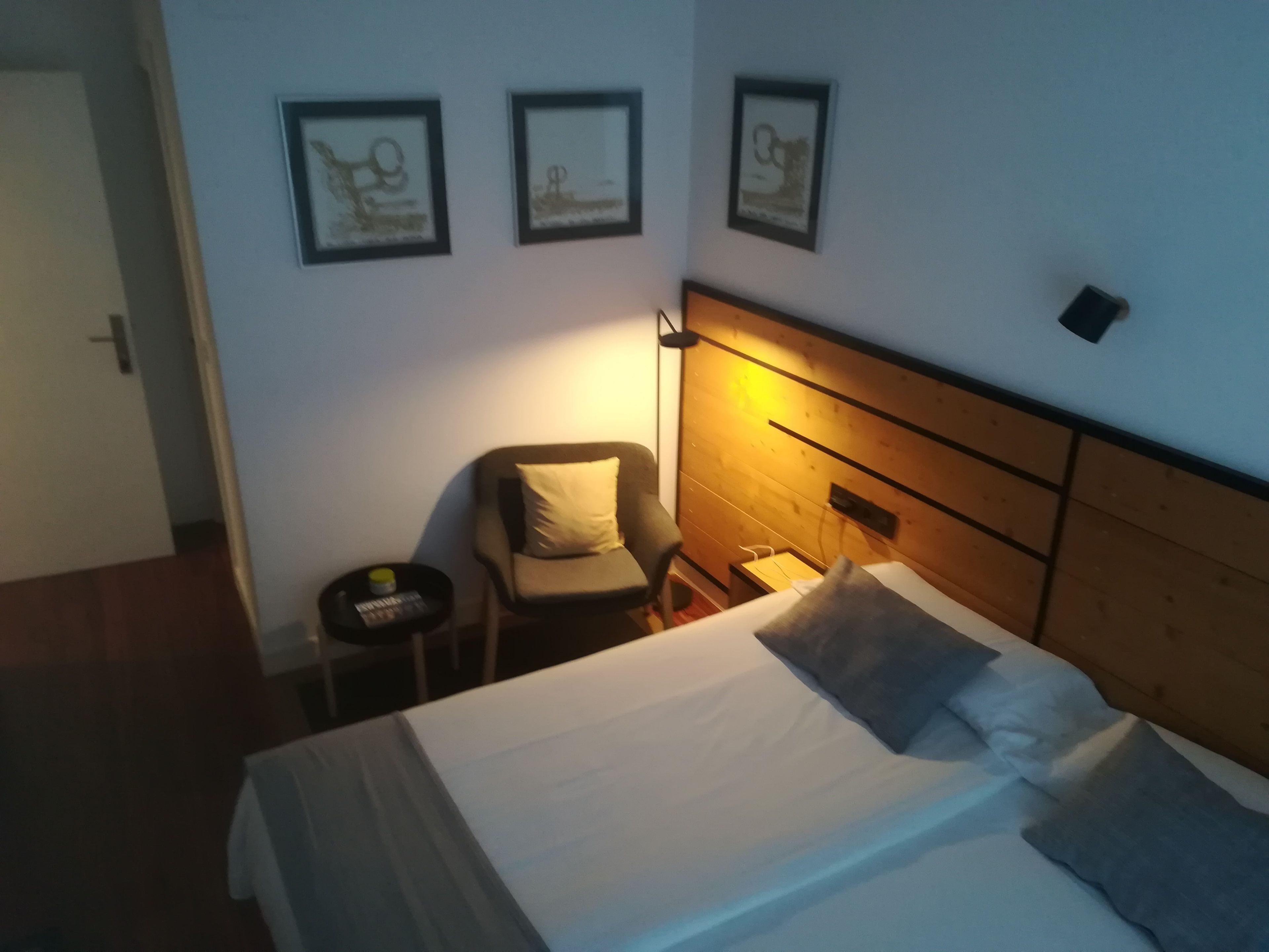 A hotel room with a bed, a chair, a nightstand, and three framed pictures on the wall.