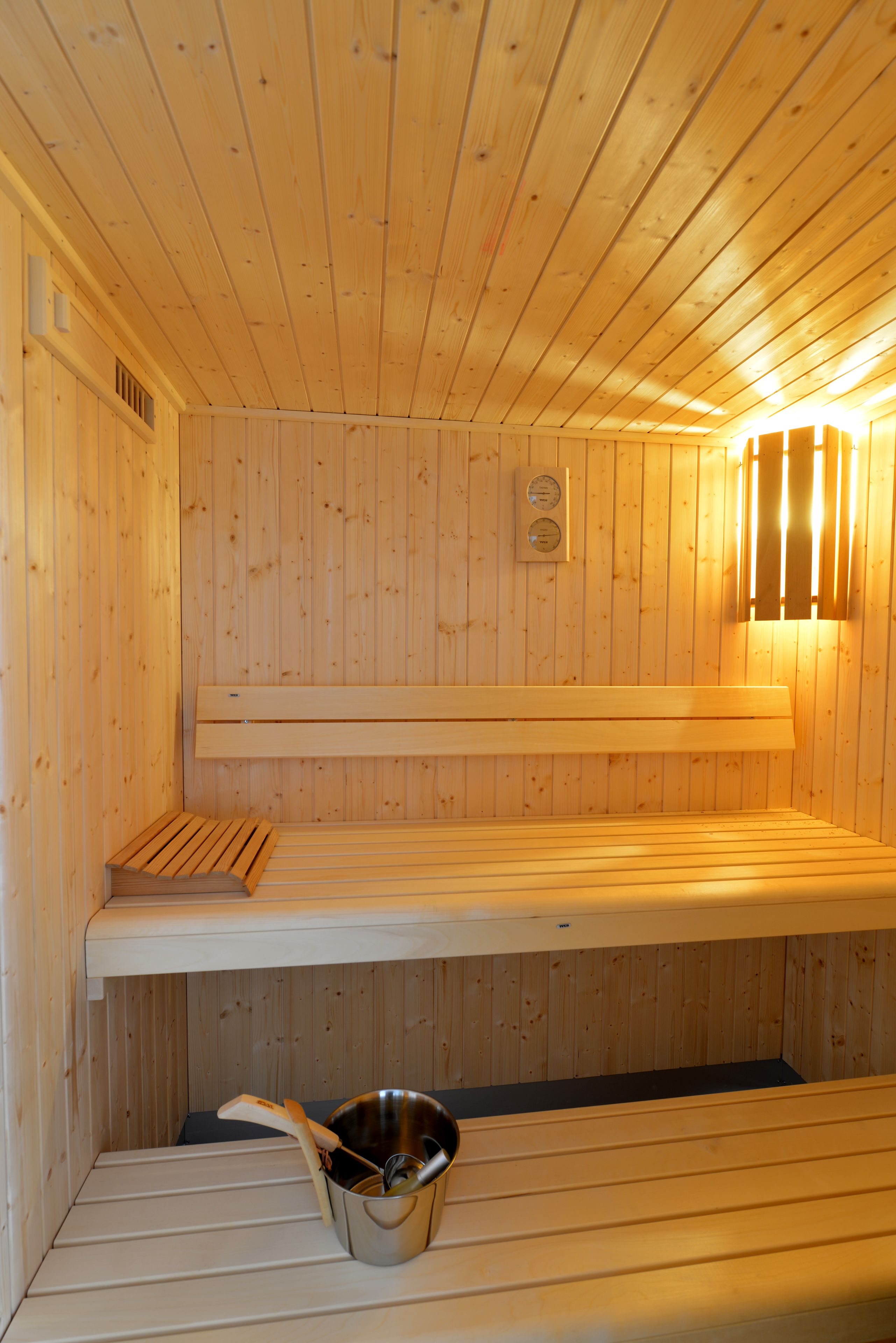 Una sauna con paredes y bancos de madera, un cubo con una cuchara, y un termómetro en la pared.