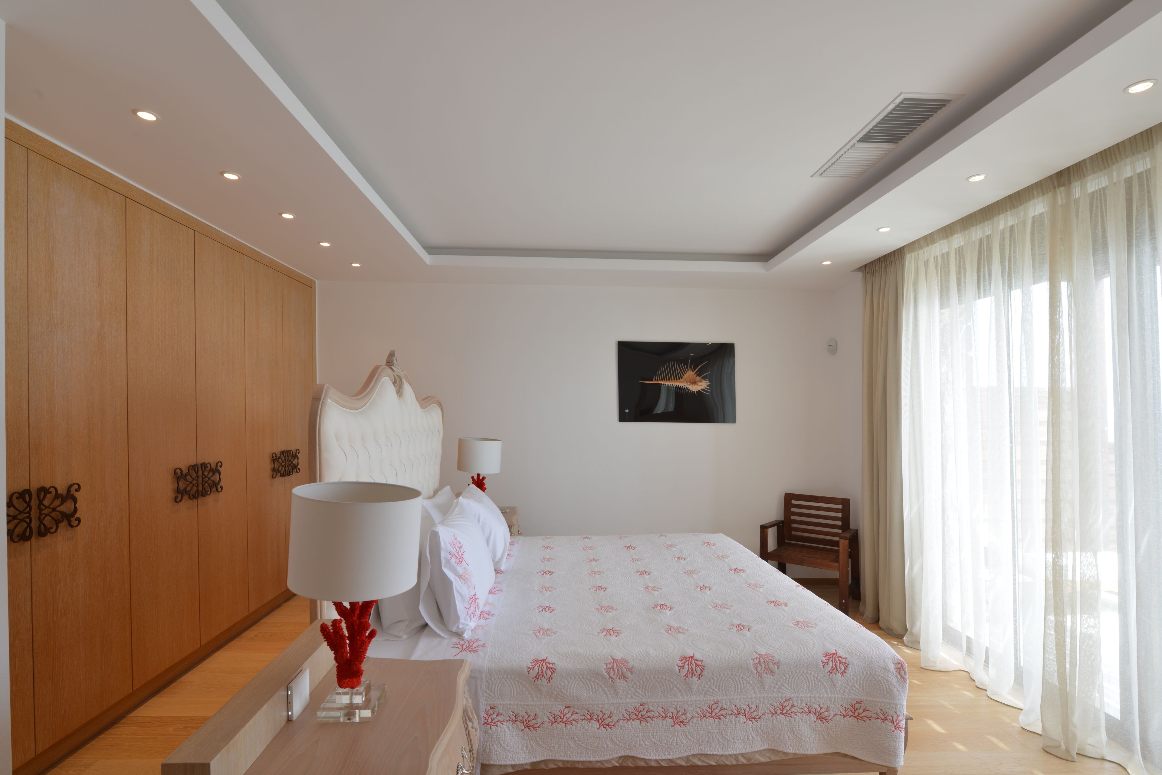 A hotel room with a bed, a wooden wardrobe, a desk with a lamp, a chair, and a window with curtains.
