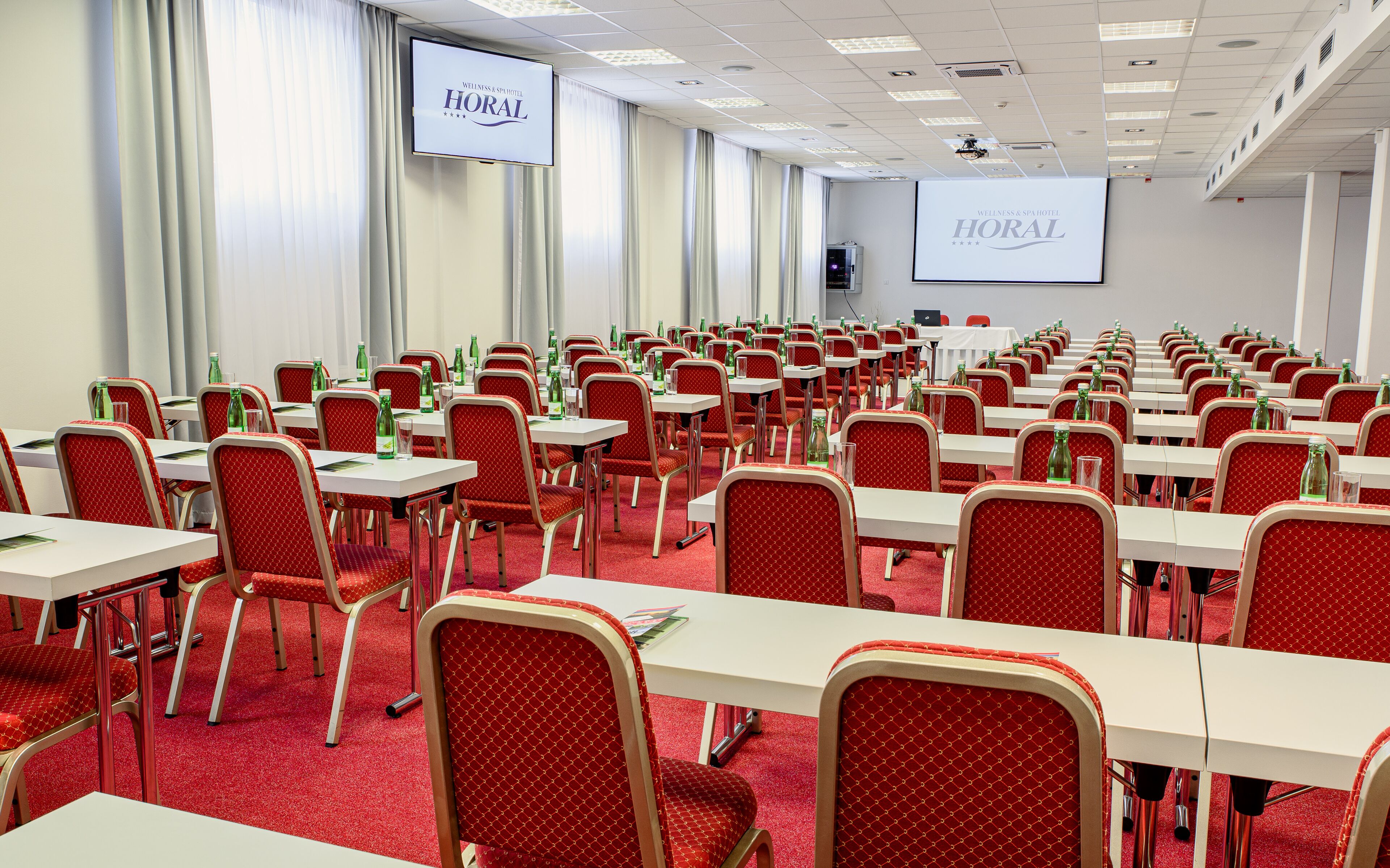 Une salle de conférence avec des rangées de chaises rouges et de tables blanches, un grand écran affichant le nom de l’hôtel, et un projecteur.