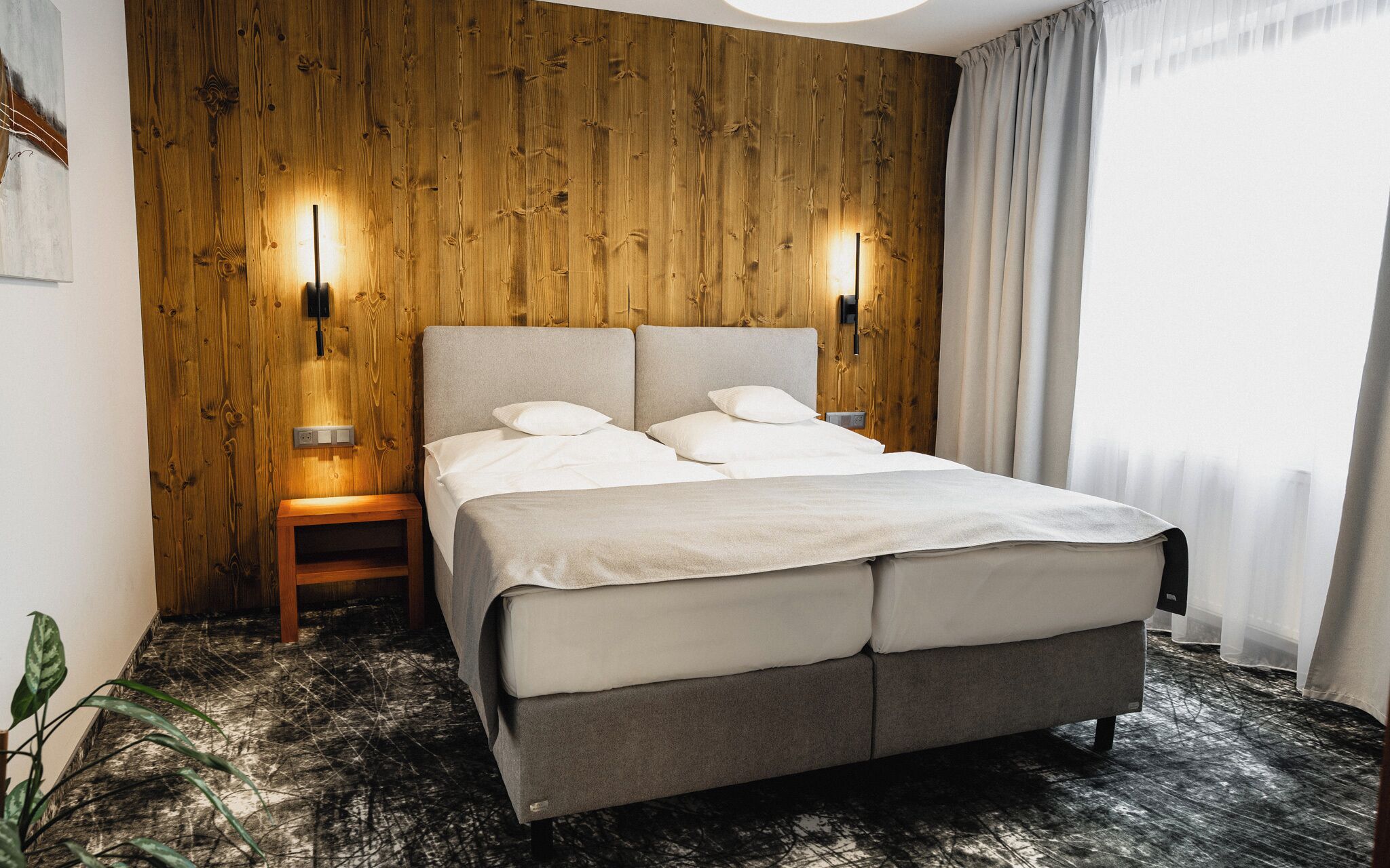 A double bed with a grey headboard and footboard, positioned against a wooden wall with two wall-mounted lights. A small wooden nightstand with a lamp is to the left of the bed.