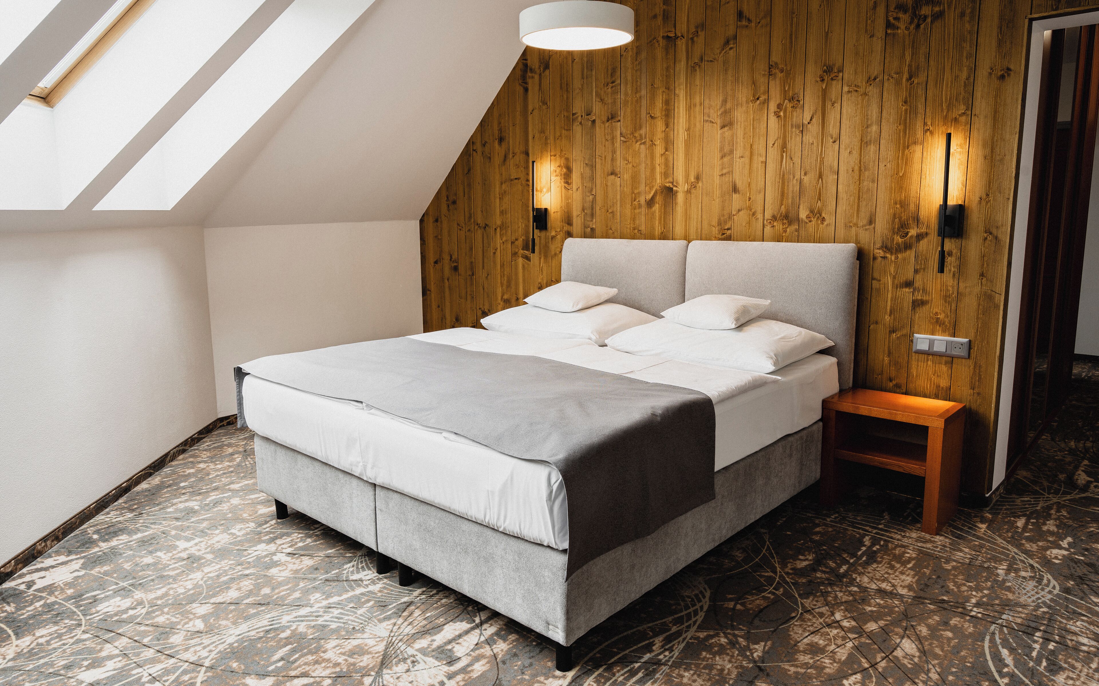 Un dormitorio moderno con techo inclinado, paneles de madera, una cama con ropa de cama gris y una mesita de noche pequeña.