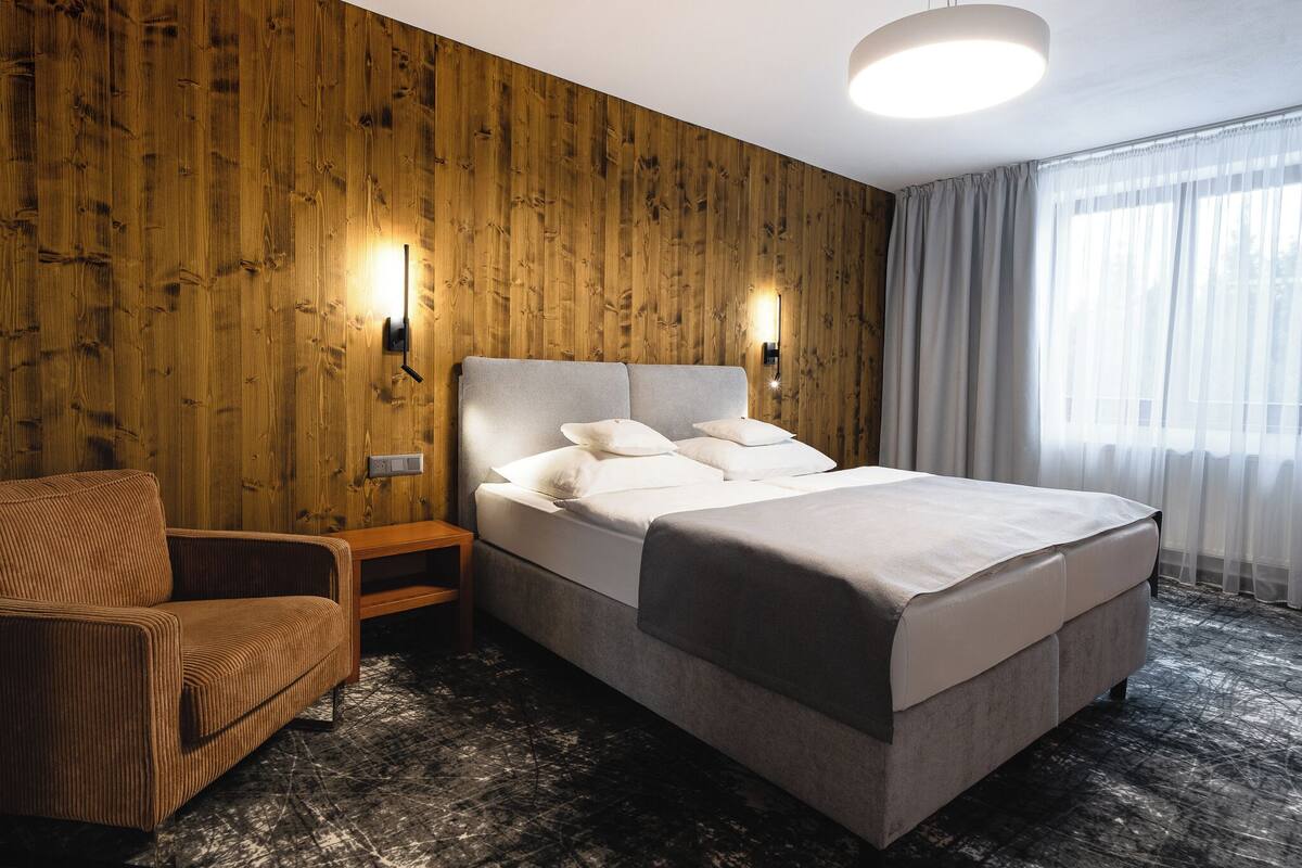 A hotel room with a wooden wall, a bed with white bedding, a brown sofa, and a nightstand.