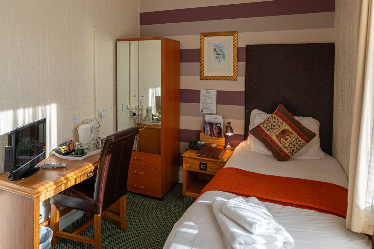 A hotel room with a bed, a desk with a computer, a chair, a wardrobe, and a bedside table with a lamp.