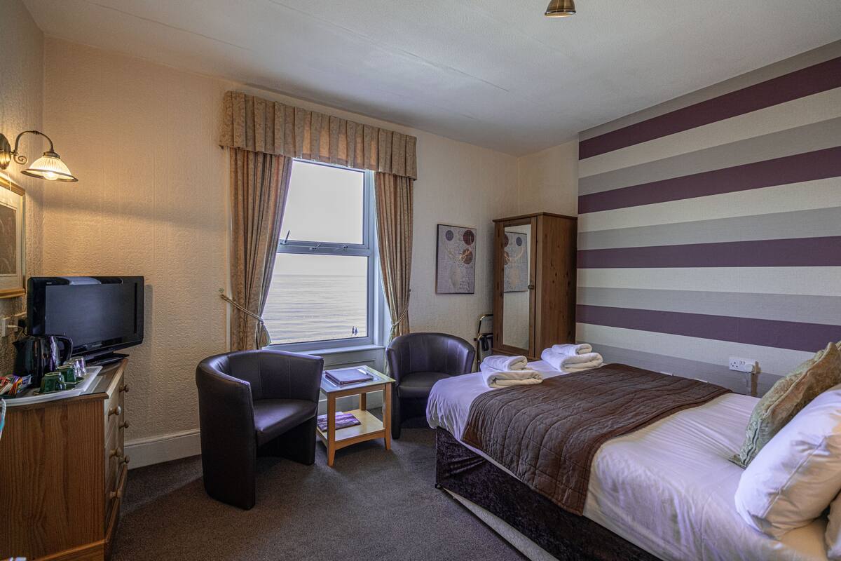 A hotel room with a bed, a desk with a television, a chair, and a window with curtains.