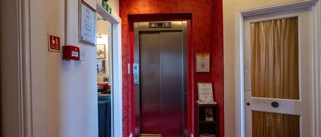 A hallway with a red carpet leading to an elevator, a door with a window, and a small shelf with items.