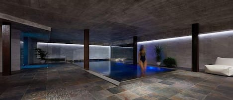 A modern indoor swimming pool with a stone floor, surrounded by walls and pillars, and a bench on one side.