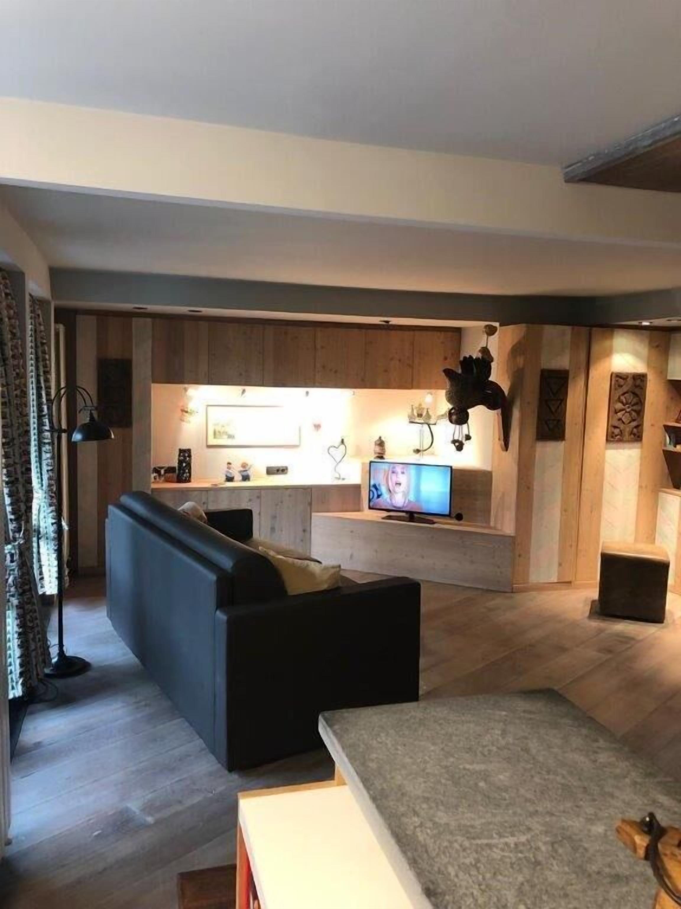 A modern living room with a sofa, a coffee table, and a TV. There is a kitchen area in the background with wooden cabinets and a countertop.