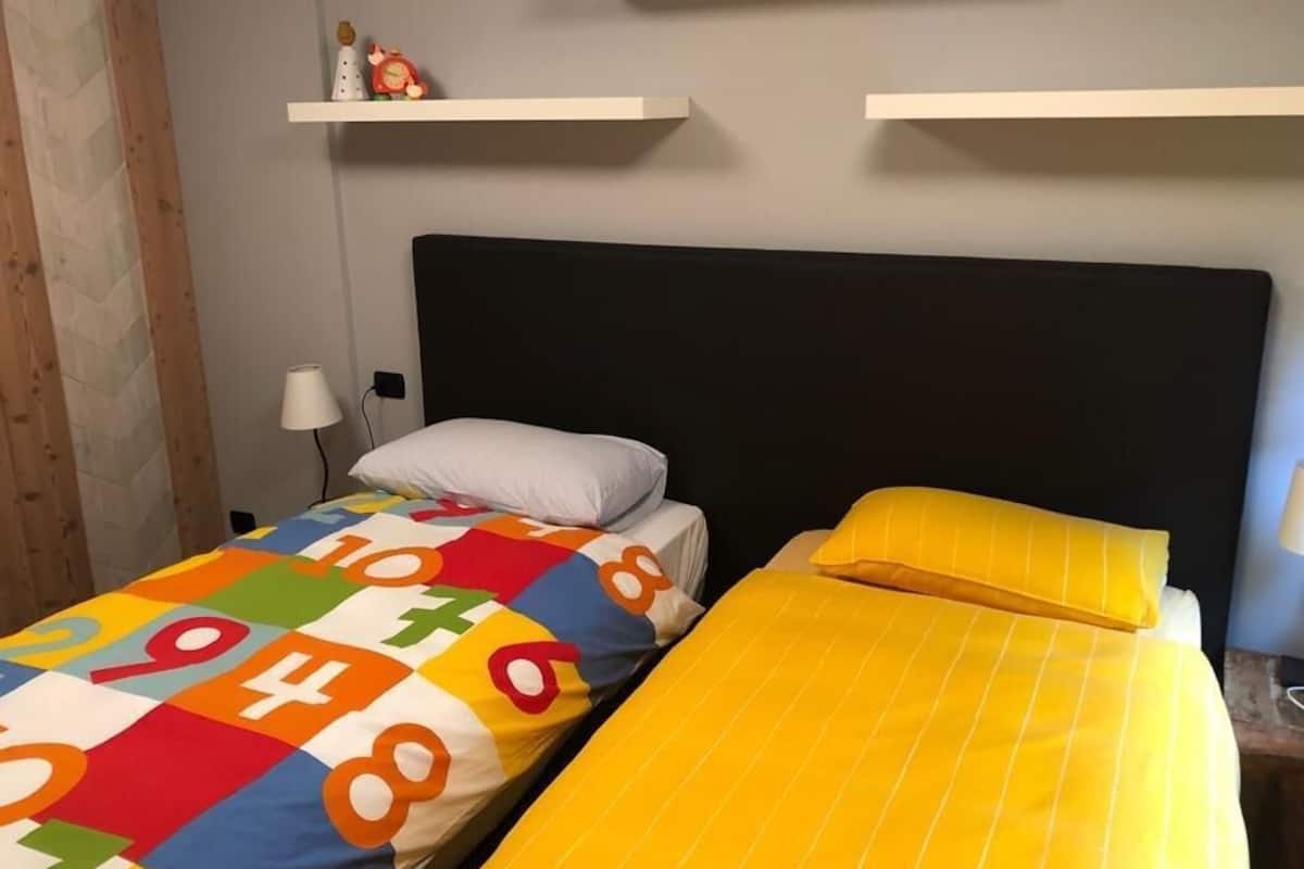 Ein Zimmer mit zwei Betten, eines mit einer farbenfrohen Steppdecke und das andere mit einer gelben Tagesdecke. Darüber befinden sich weiße Regale und ein kleiner Tisch mit einer Lampe.