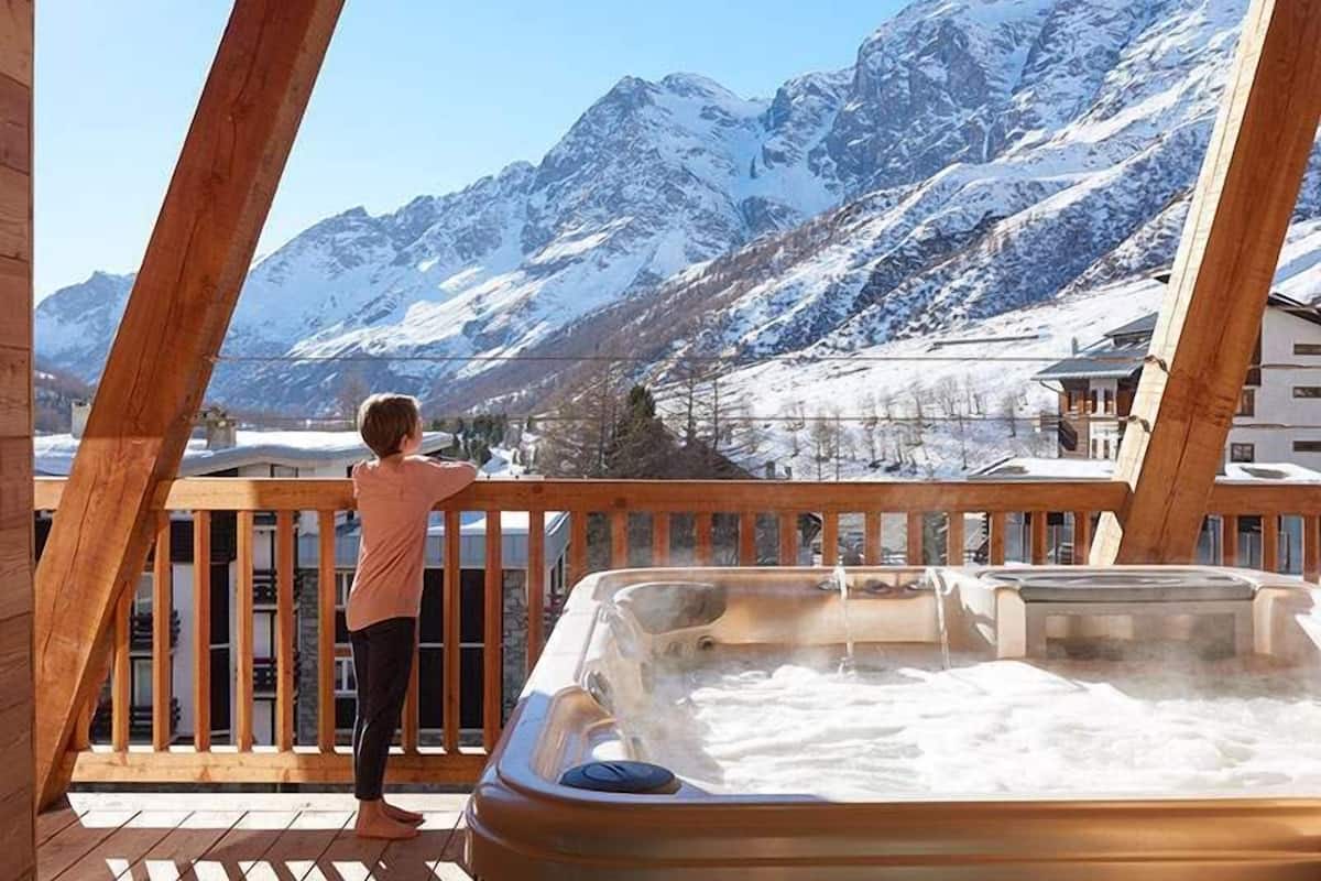 Eine Person steht auf einer Holzterrasse mit Blick auf eine gebirgige Landschaft. Im Vordergrund befindet sich eine Whirlwanne.