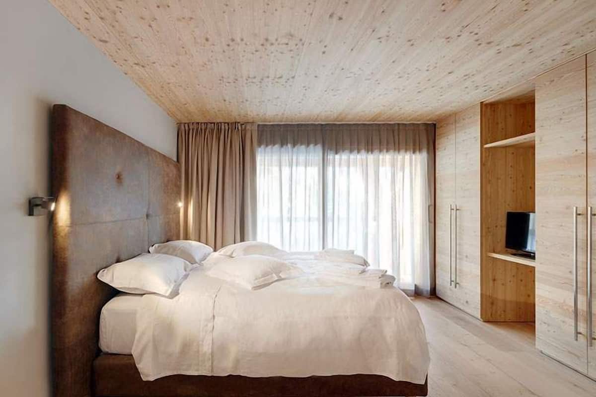 Ein Schlafzimmer mit Holzboden, einem Bett mit weißen Bettwäsche, einem Fernseher auf einer Holzablage und einem großen Fenster mit Vorhängen.