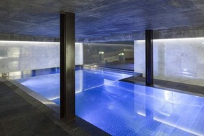 A modern indoor swimming pool with a blue-lit floor, surrounded by walls with a textured finish. - Aux Pieds du Roi (Valtournenche)