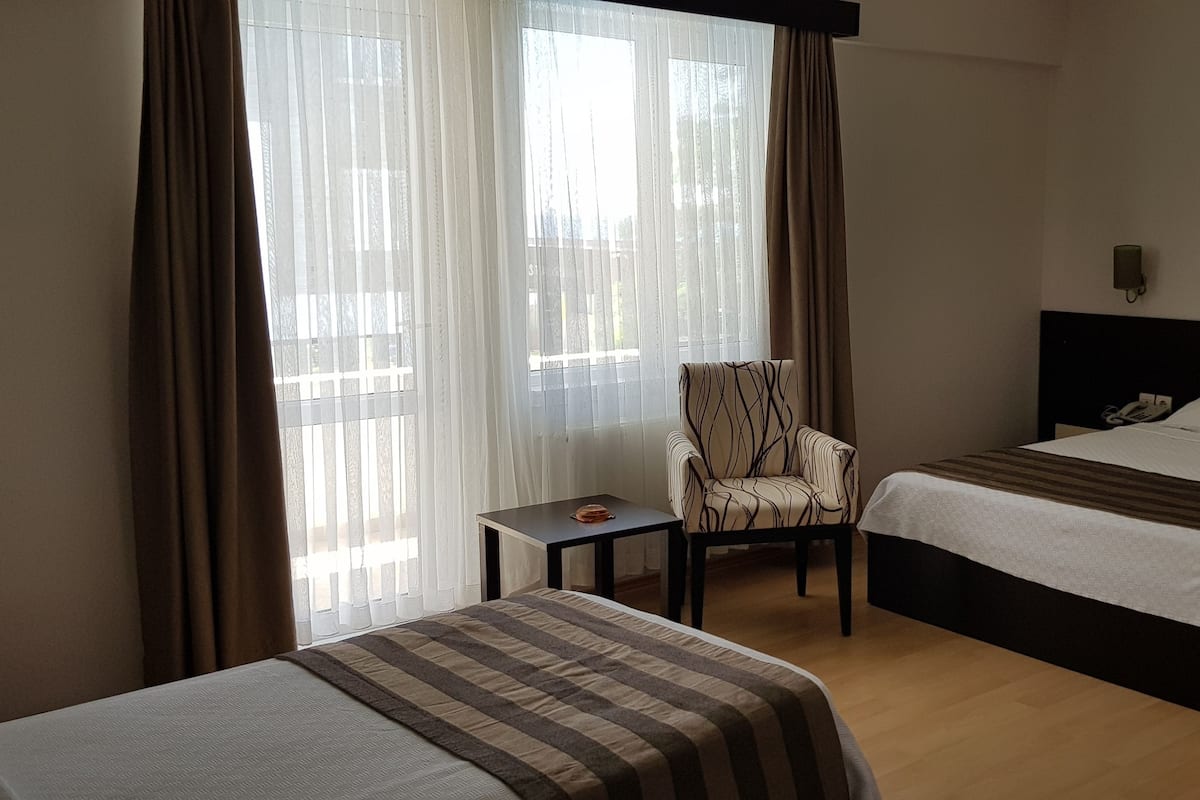 Une chambre d’hôtel avec deux lits, une chaise, une petite table et des rideaux légers.