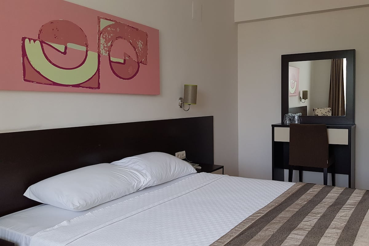 Une chambre d’hôtel équipée d’un lit, d’un bureau, d’une chaise, d’un miroir et d’une lampe fixée au mur.