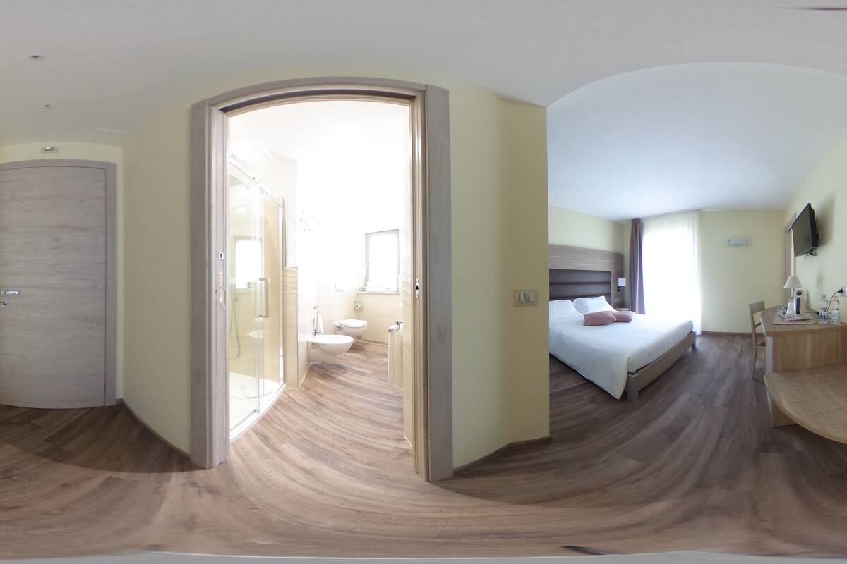 A modern hotel room with a wooden floor, a bed with a headboard, a bedside table, a TV mounted on the wall, and a bathroom with a toilet and sink.