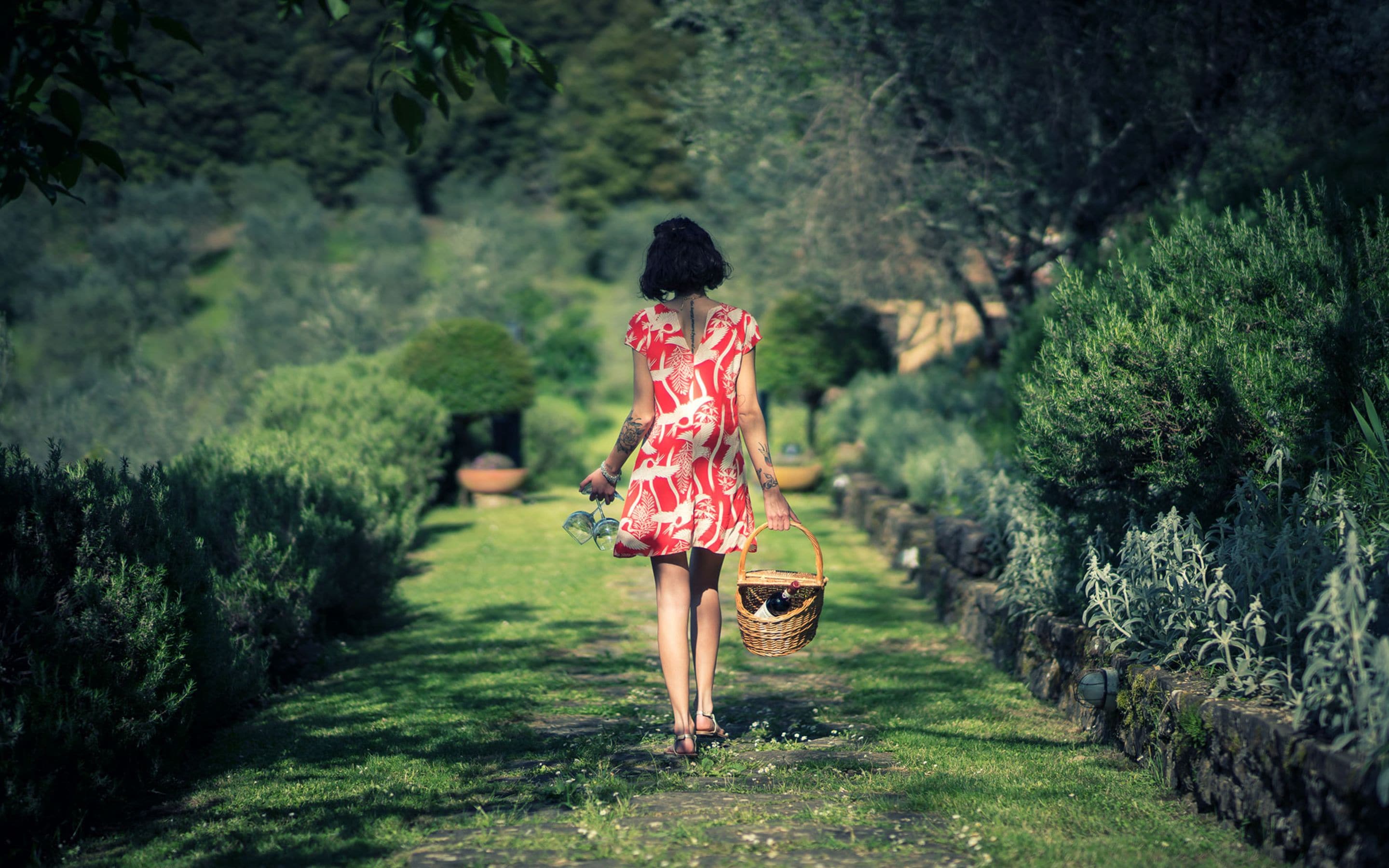 A woman walking down a garden path with a basket, surrounded by greenery and stone walls.
