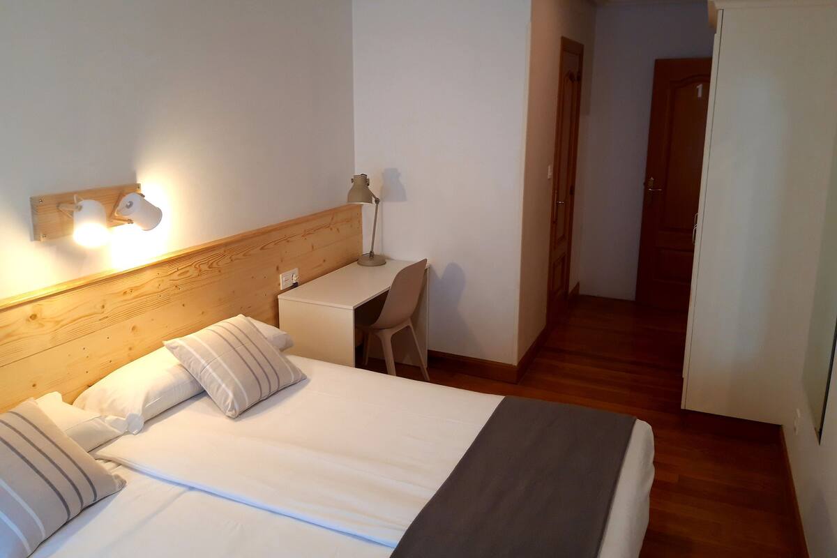 A hotel room with a bed, bedside table, lamp, and a desk with a chair.