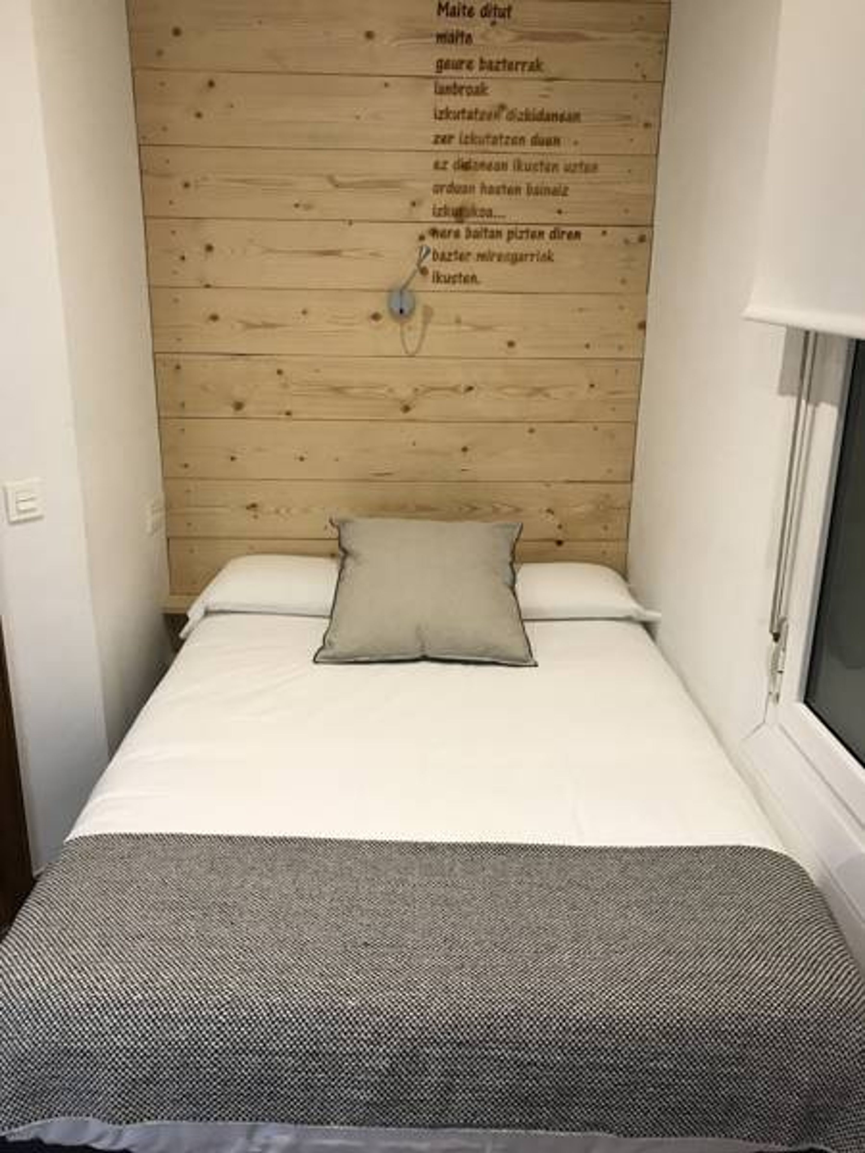Habitació petita amb un llit individual, un panell de fusta al capçal i una finestra.