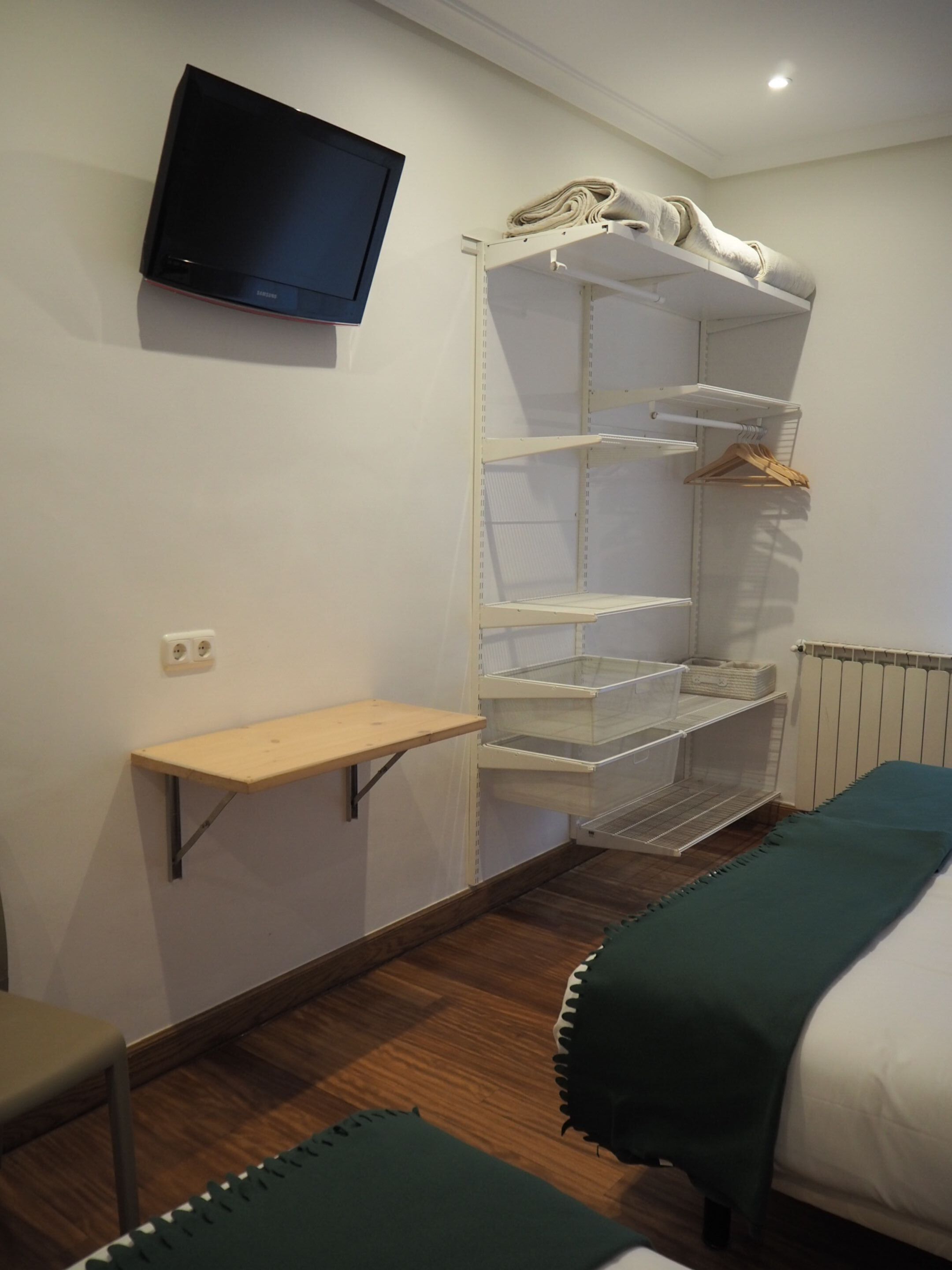 Habitación de hotel con televisor montado en la pared, armario empotrado con estantes, una pequeña mesa de madera y dos camas con cubrecamas verdes.