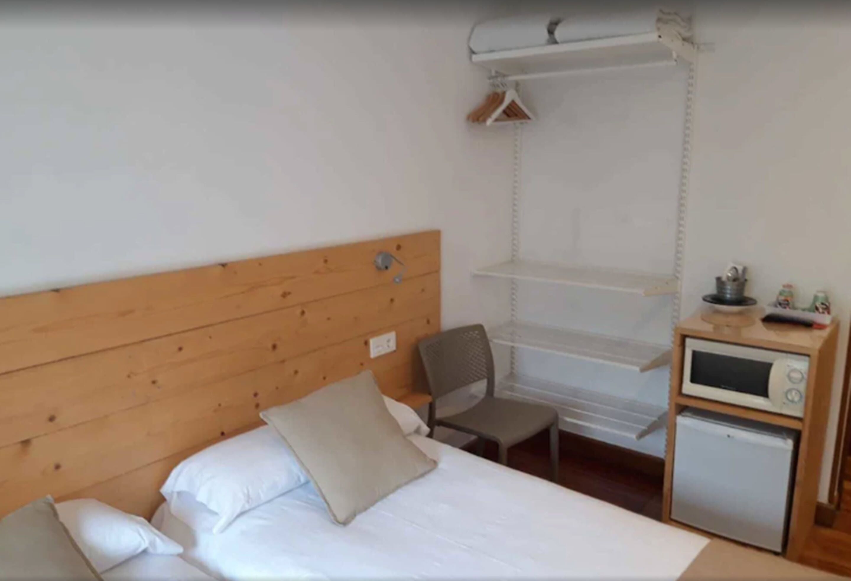 A bedroom with a wooden headboard, a bed with white bedding, a small desk with a chair, a microwave, and a mini-fridge.