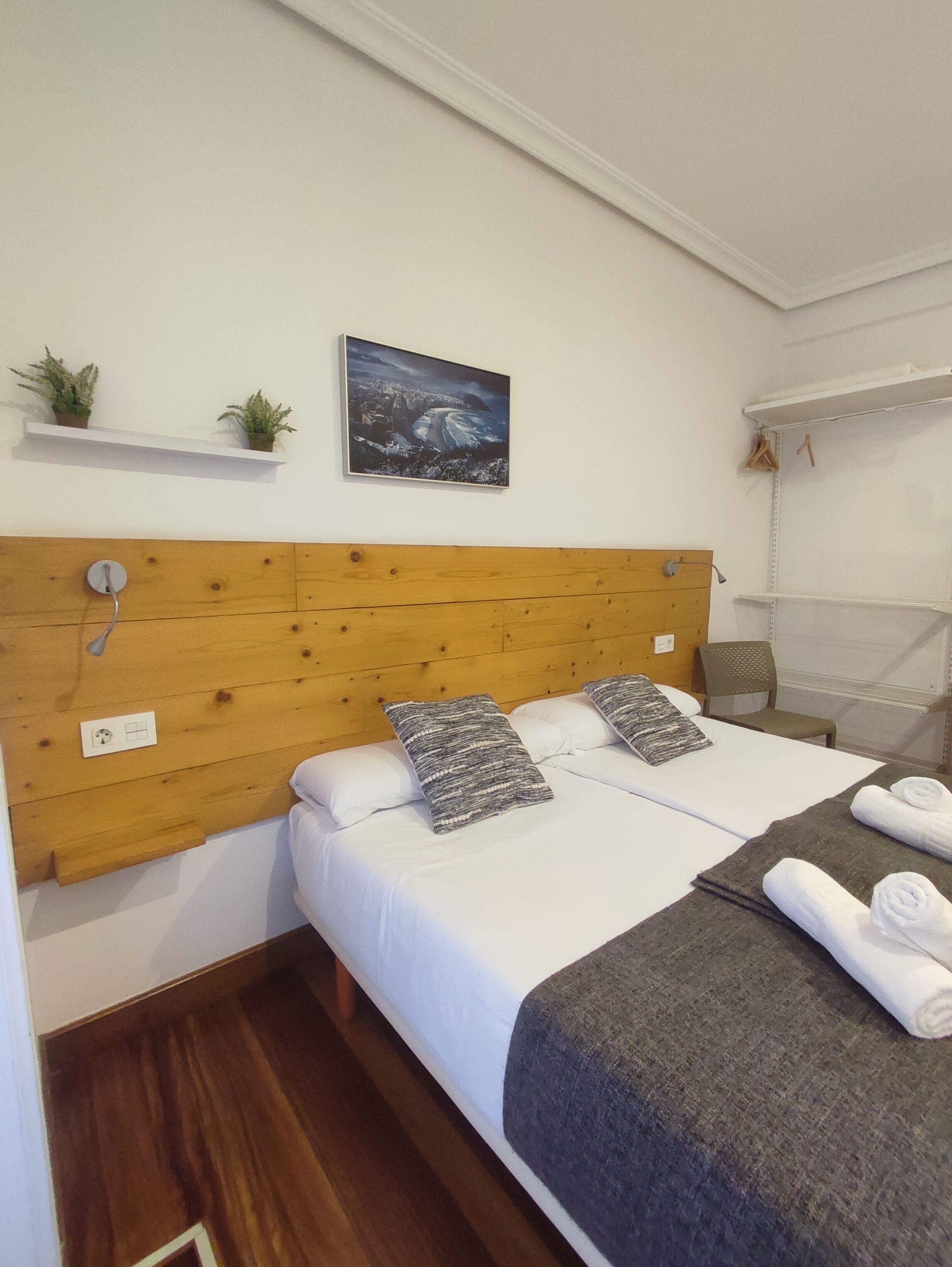 A double bed with white linens, two pillows, a gray throw blanket, and rolled towels on a wooden headboard. A small shelf with two potted plants and a framed picture of a landscape are on the wall above the bed.