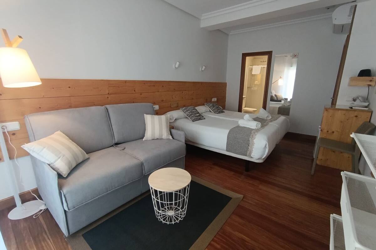 A modern room with a grey sofa, a small round table, a bed with a wooden headboard, and a mirror on the wall.