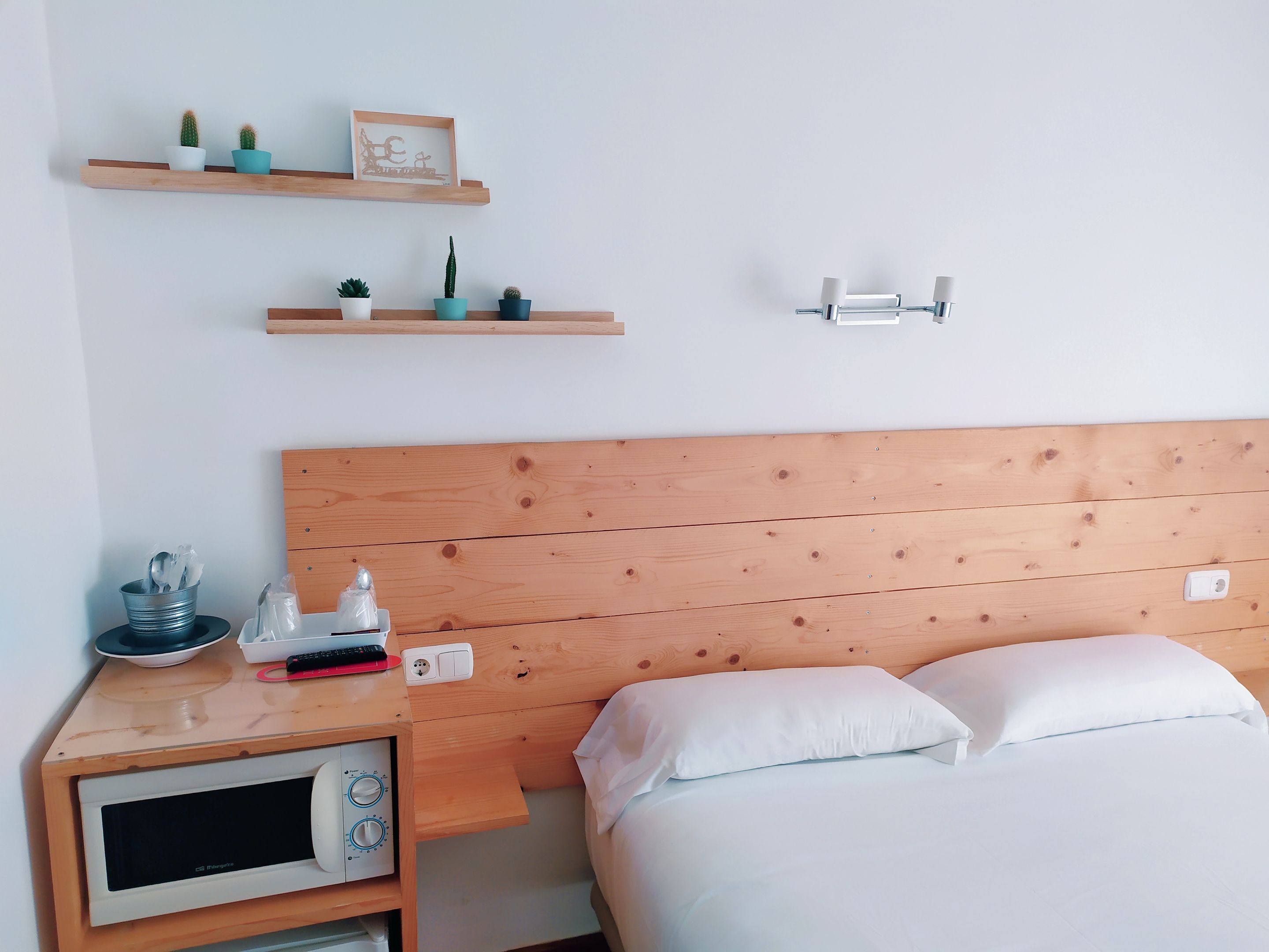 A bedroom with a wooden headboard, a bed with white linens, a nightstand with a microwave, and two wall-mounted shelves with decorative items.