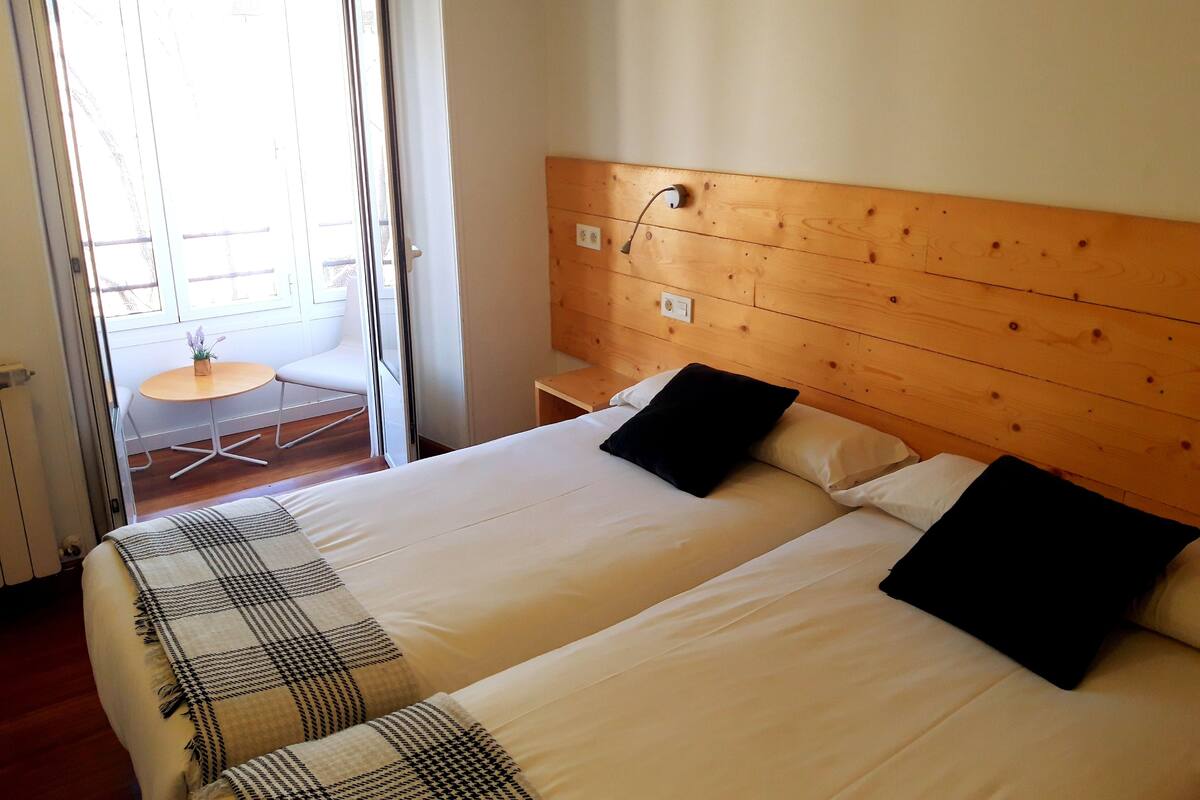 A hotel room with two beds, a wooden headboard, a small table with a chair, and a view through a glass door.
