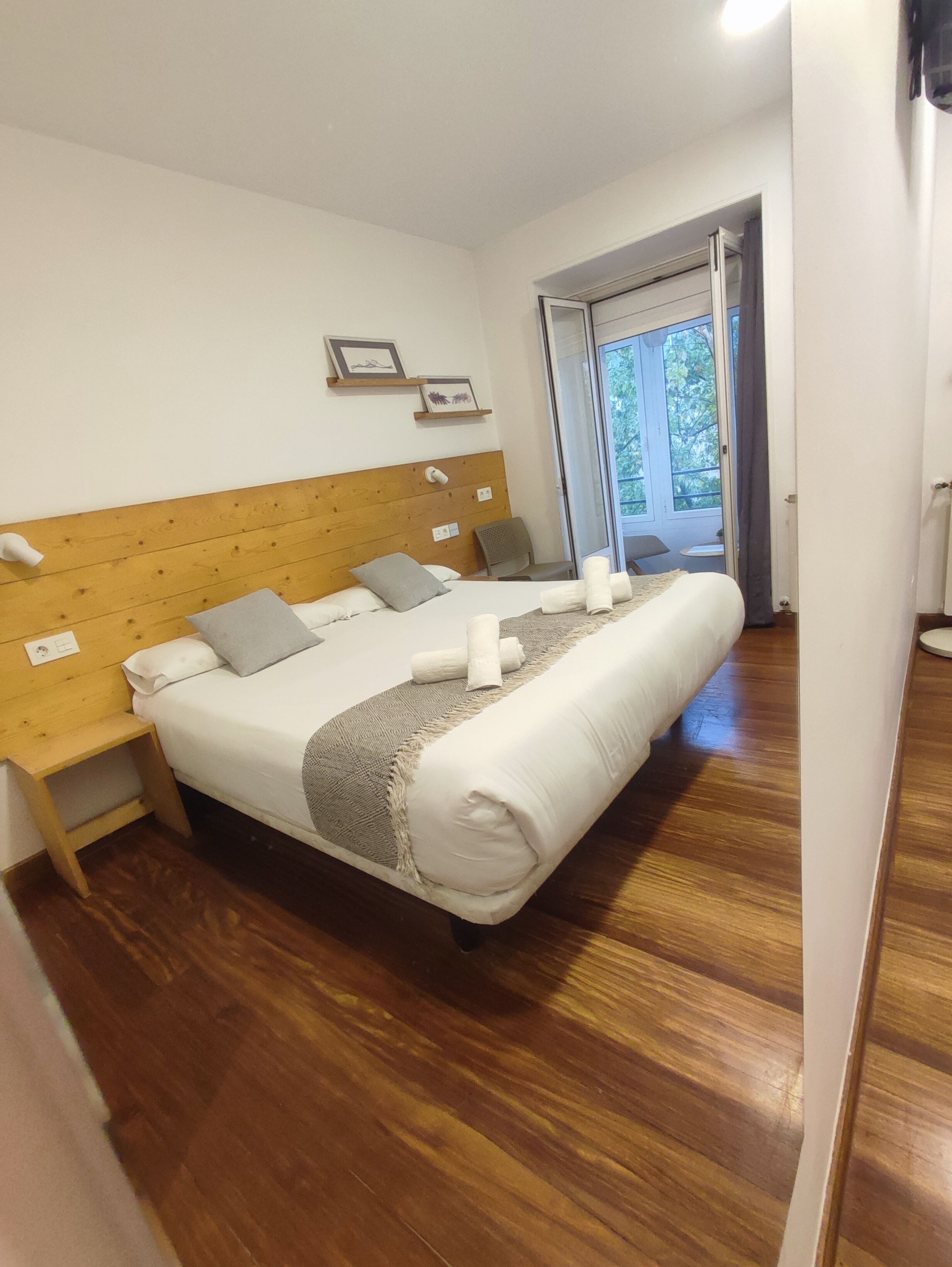 Un dormitorio con cabecera de madera, cama con sábanas blancas, dos mesitas de noche y vistas al exterior a través de una puerta.