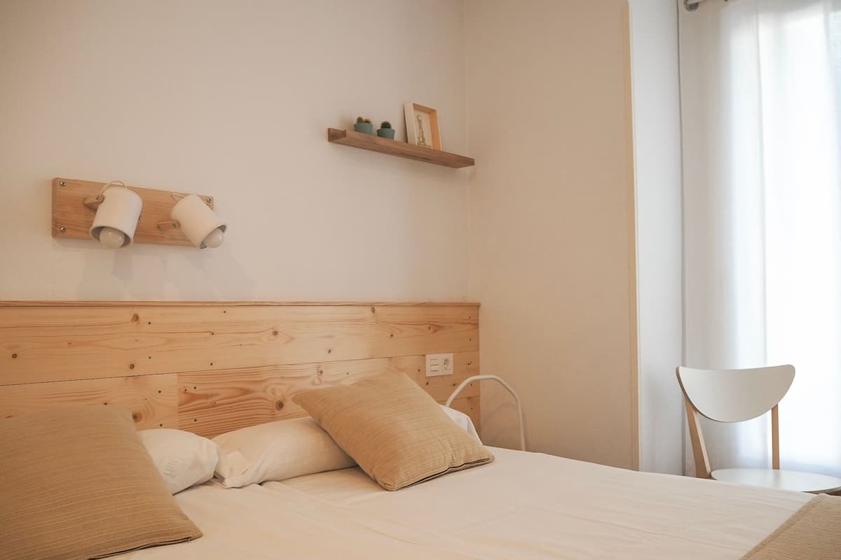 A bedroom with a wooden headboard, a bed with beige bedding, two pillows, a bedside table, a chair, and a shelf with decorative items.