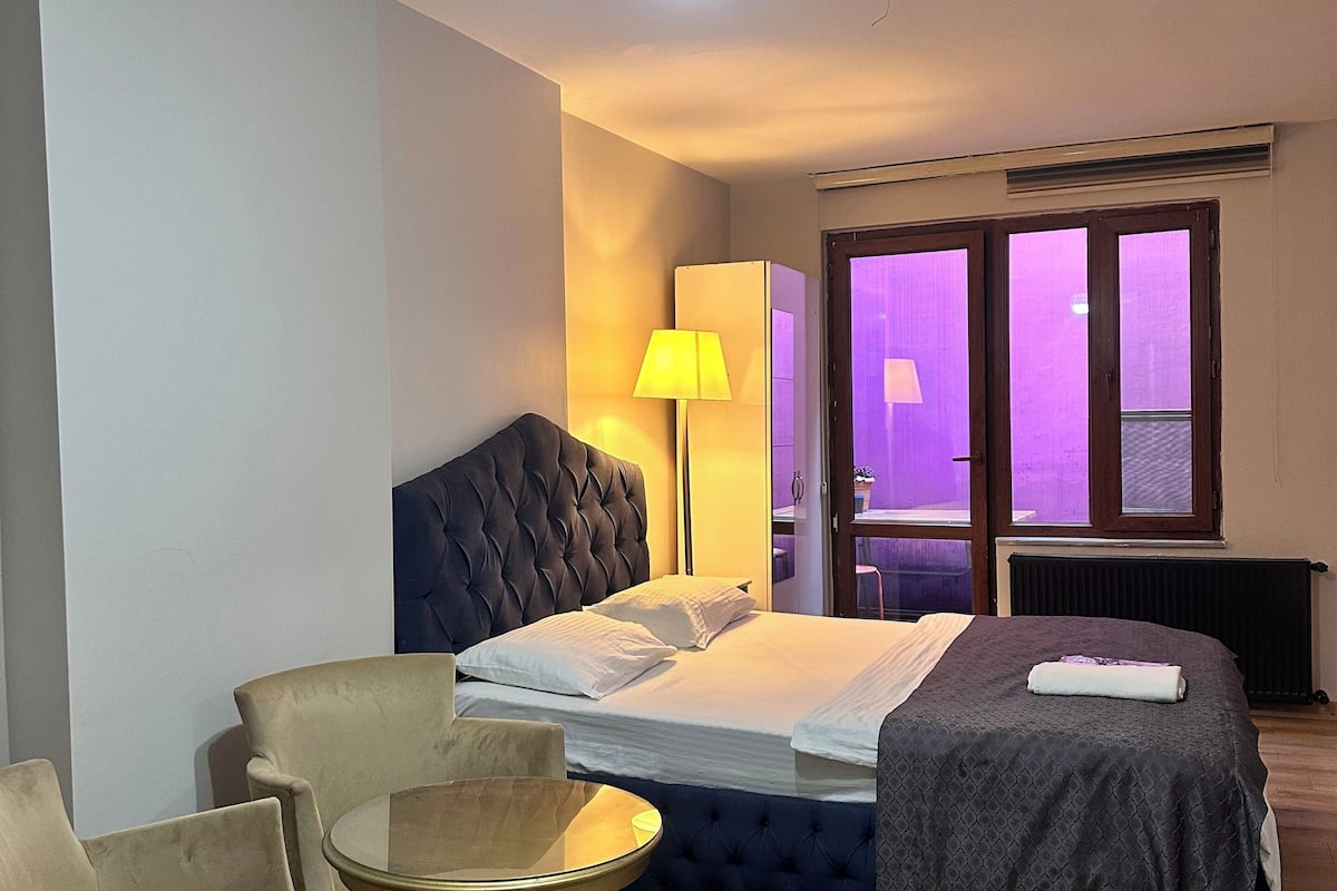 A hotel room with a bed, a round table, two armchairs, a lamp, and a ceiling light fixture.