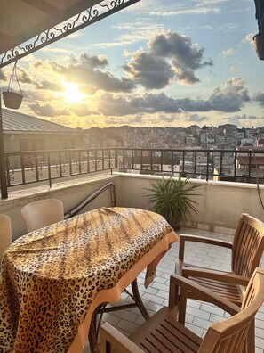A balcony with a table covered in a leopard print tablecloth, surrounded by wooden chairs, overlooking a cityscape with buildings and a sunset. - Centre Suites Taksim (Istanbul)