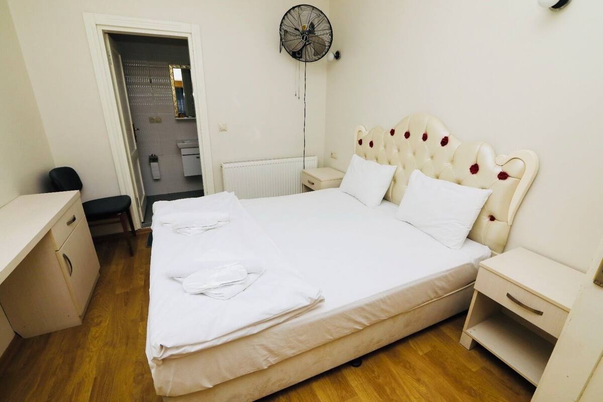 A neatly made bed with white linens, a bedside table, a chair, and a wall-mounted fan.
