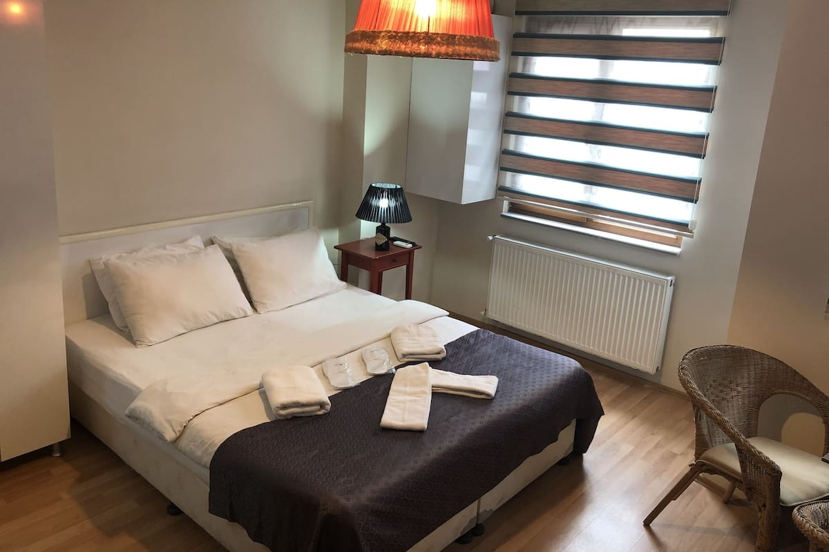 A neatly made bed with white and dark linens, a bedside lamp, and a chair in a room with wooden flooring and a window with blinds.