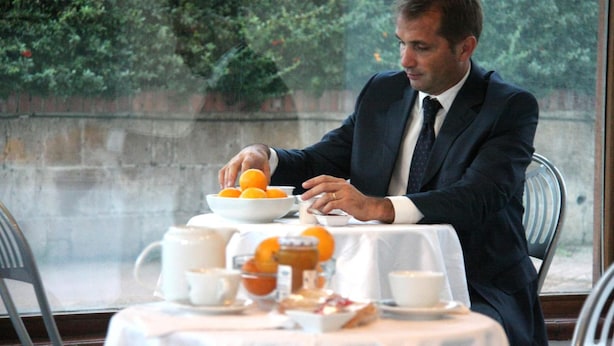 Een man in een donker pak zittend aan een tafel met een witte tafelkleed, een schaal oranjes en diverse ontbijtartikelen.