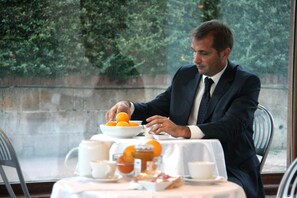 Un homme en costume sombre est assis à une table recouverte d’une nappe blanche. On y voit un bol d’oranges et divers aliments pour le petit-déjeuner.