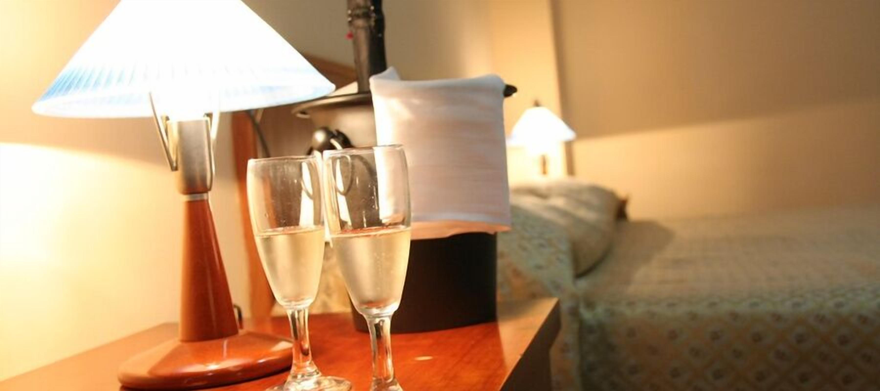 A wooden nightstand with two glasses of water, a lamp, and a tissue box in a hotel room.
