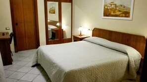 A hotel room with a bed, a wooden headboard, a bedside table with a lamp, a mirror, and a door. - Hotel Il Ciliegio (Santo Stefano del Sole)