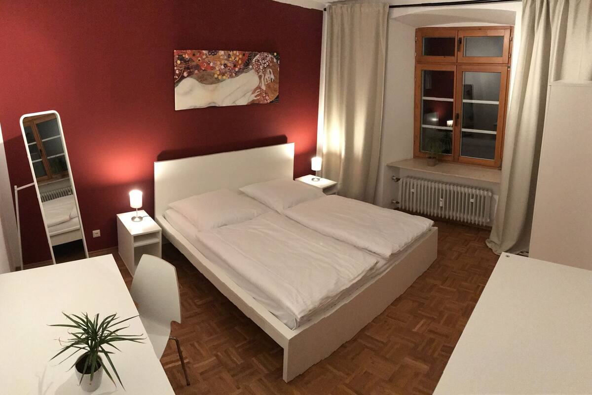 A bedroom with a bed, bedside tables, a desk, a chair, a potted plant, a window with curtains, a radiator, and a painting on the wall.