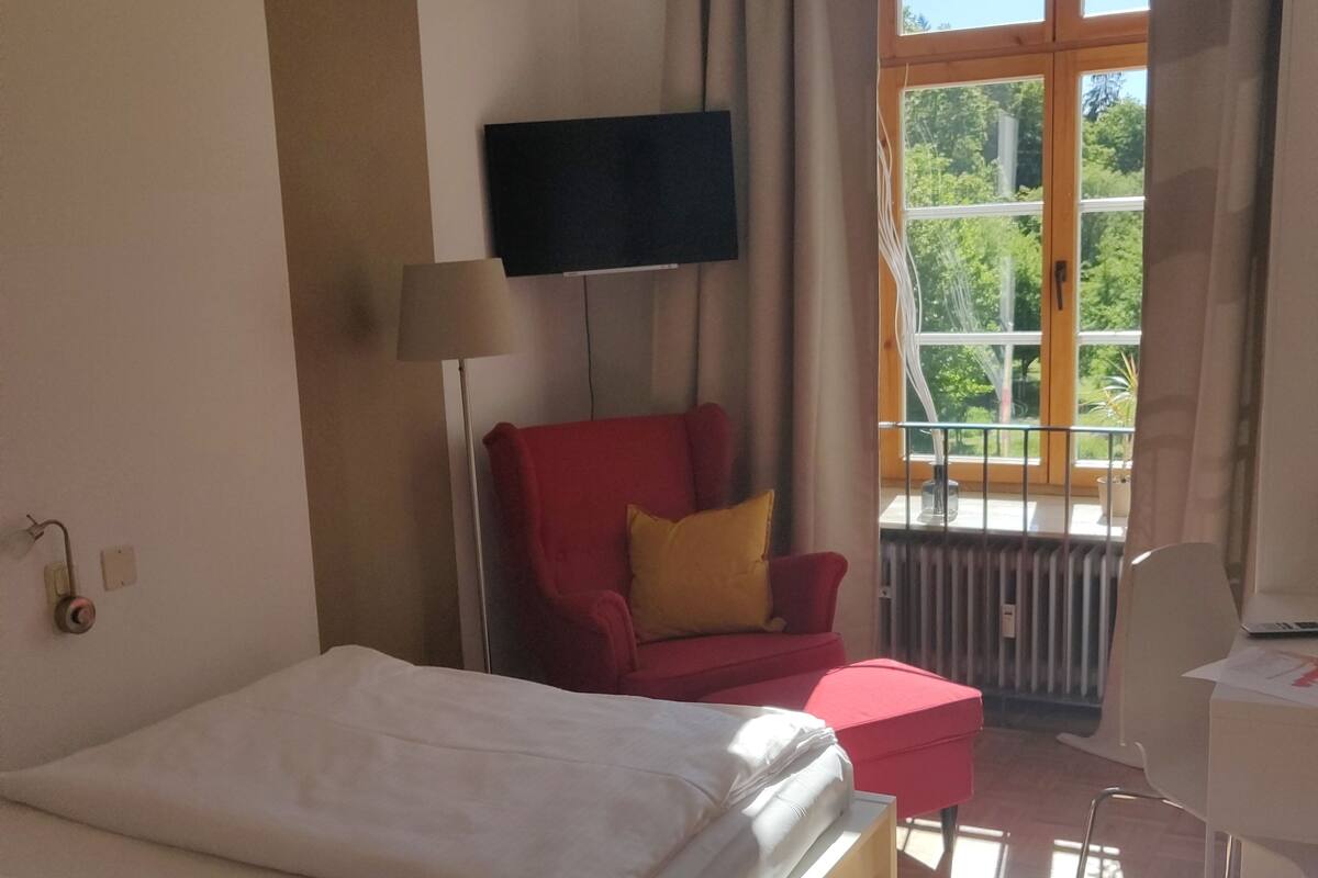 A hotel room with a bed, a red armchair, a small table, a TV, and a window with a view of greenery.