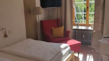 A hotel room with a bed, a red armchair, a small table, a TV, and a window with a view of greenery.
