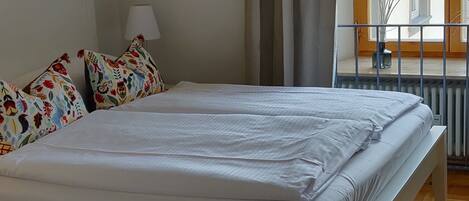 Un dormitorio con una cama grande, una lámpara negra colgante, una ventana con cortinas y suelo de madera.