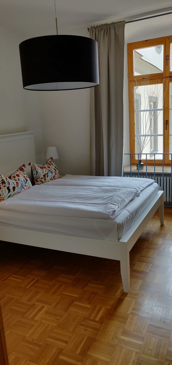 A bedroom with a large bed, a hanging black lamp, a window with curtains, and a wooden floor. - Gästehaus Abtei Sayn (Bendorf)