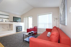 A modern living room with a red sofa, a glass coffee table, a flat-screen TV mounted on the wall, a fireplace, and a large window with blinds. - Pelicanstay in Churchill Meadows (Mississauga)