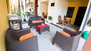 A covered outdoor seating area with wicker furniture and a small table, surrounded by potted plants and a staircase leading to an upper level. - Papaya'S Lodge (Tamanique)